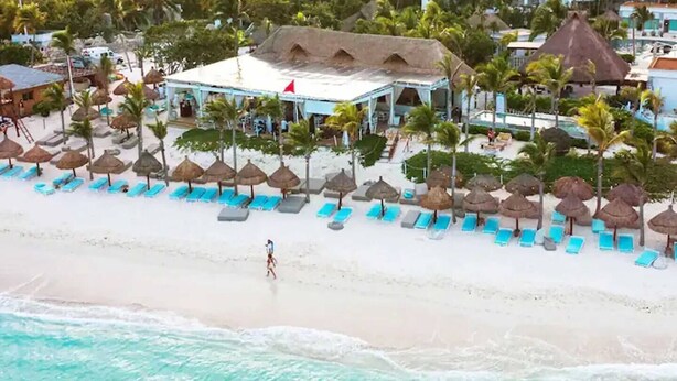 Um resort à beira-mar com uma fileira de espreguiçadeiras sob guarda-sóis de palha, um edifício com uma bandeira vermelha e uma pessoa caminhando na praia.