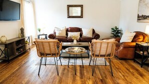 Een woonkamer met een bruine leren bank, twee rieten fauteuils, een glazen salontafel en een aan de muur bevestigde televisie.