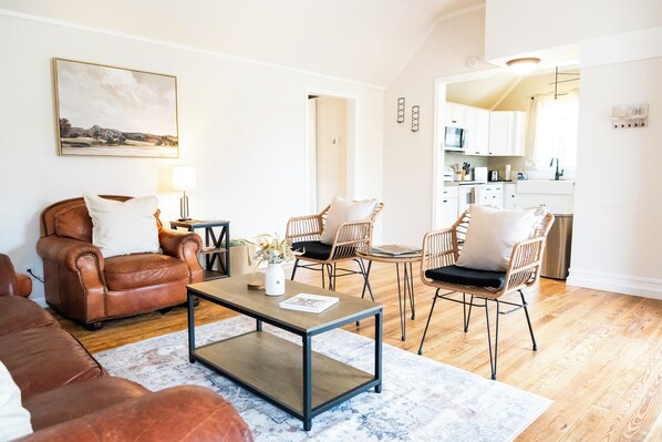A living room with a brown leather sofa, a coffee table, and wicker chairs. There is a painting on the wall and a kitchen area in the background. - Charming Getaway in Comfort, TX - Relax, Recharge, Explore - Texas Wine Country (Comfort)