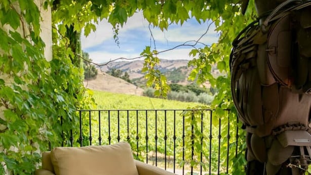 A beige sofa under a vine-covered pergola with a view of a vineyard and hills in the background. - The Vineyard House in the Pastures of Heaven (Corral de Tierra)