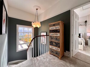 A staircase with a railing, a wooden cabinet with drawers, a window with a view of trees, and a door leading to another room. - Beautiful 5-Bed Regency Home in Montpellier (Gloucestershire)