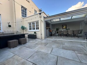 A patio area with a dining table and chairs, a black outdoor kitchen, and a glass door leading to an interior space. - Beautiful 5-Bed Regency Home in Montpellier (Gloucestershire)