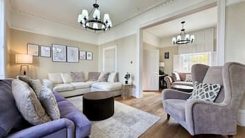 Een ruime woonkamer met een grote bank, een kleinere fauteuil en een salontafel. Twee kroonluchters hangen aan het plafond.