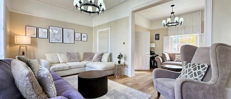 A spacious living room with a large sofa, a smaller armchair, and a coffee table. There are two chandeliers hanging from the ceiling.