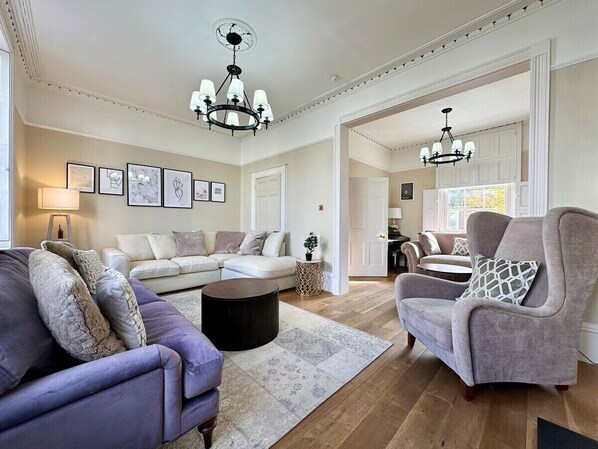 Un salon spacieux avec un grand canapé, un fauteuil plus petit et une table basse. Deux lustres sont suspendus au plafond.