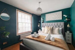 Une chambre avec un grand lit, une table de chevet, une commode, un luminaire suspendu et une fenêtre avec des stores.