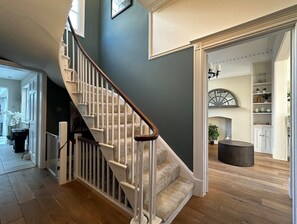 Un escalier avec des rampes blanches et des marches recouvertes d’une moquette beige, menant à une pièce dotée d’un plancher en bois et d’un pouf rond noir.