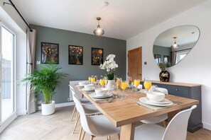 A dining area with a wooden table set for six, white chairs, and a large mirror on the wall. - Comfortable & Contemporary 5-Bed 3 Bathroom Home (Gloucestershire)