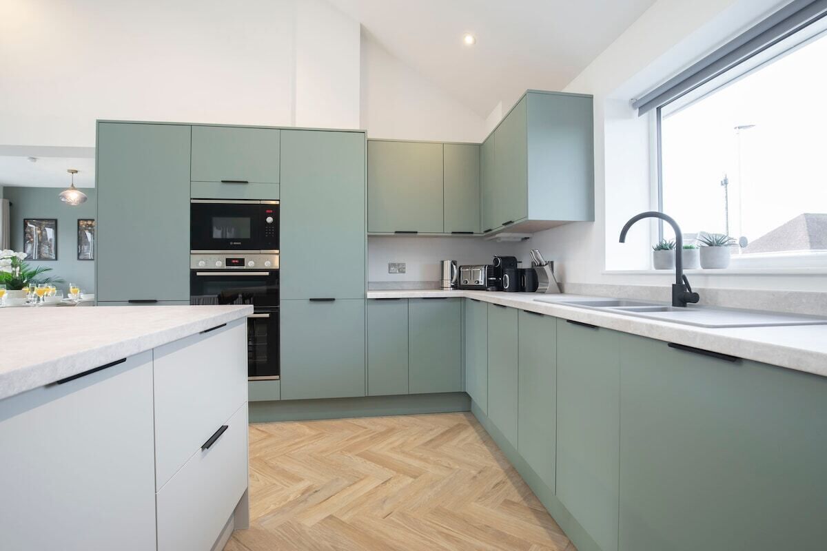 A modern kitchen with light green cabinets, a built-in oven, and a large window providing natural light.