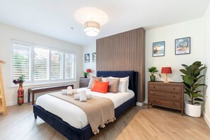 A modern bedroom with a large bed, bedside tables, a dresser, and a window with blinds. - Comfortable & Contemporary 5-Bed 3 Bathroom Home (Gloucestershire)
