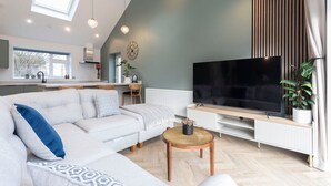 Una sala moderna con un televisor de pantalla plana grande, un sofá blanco, una mesa de centro y una cocina al fondo.