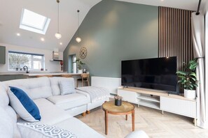 A modern living room with a large flat-screen TV, a white sofa, a coffee table, and a kitchen in the background. - Comfortable & Contemporary 5-Bed 3 Bathroom Home (Gloucestershire)