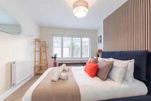 Ein Schlafzimmer mit einem Bett, einer Holzleiter, einem Heizkörper, einem Fenster mit Jalousien und einer Wand mit vertikalen Holzlatten.