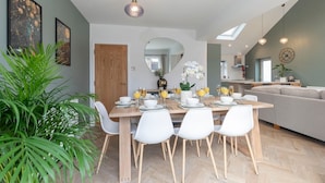 Un moderno comedor con una mesa de madera preparada para cuatro personas, rodeada de sillas blancas. Un espejo grande cuelga de la pared, y hay dos plantas en macetas.