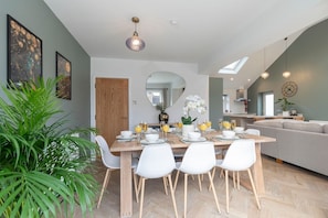 A modern dining area with a wooden table set for four, surrounded by white chairs. A large mirror hangs on the wall, and there are two potted plants.