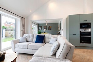 A modern living room with a beige sectional sofa, a dining table, and a kitchen area with grey cabinets and a built-in oven. - Comfortable & Contemporary 5-Bed 3 Bathroom Home (Gloucestershire)