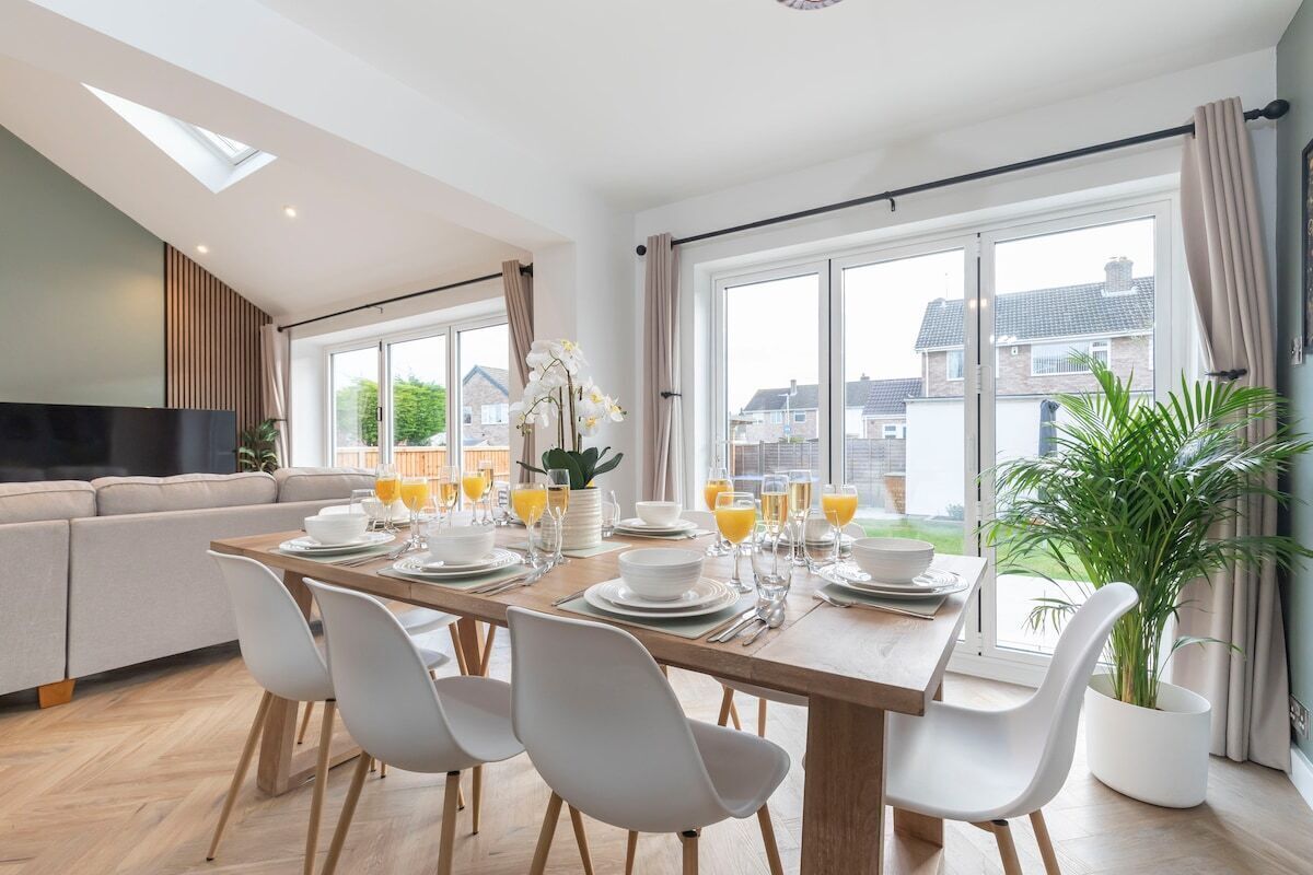 A modern dining area with a long table set for six, surrounded by white chairs. A large window offers a view of the neighborhood.