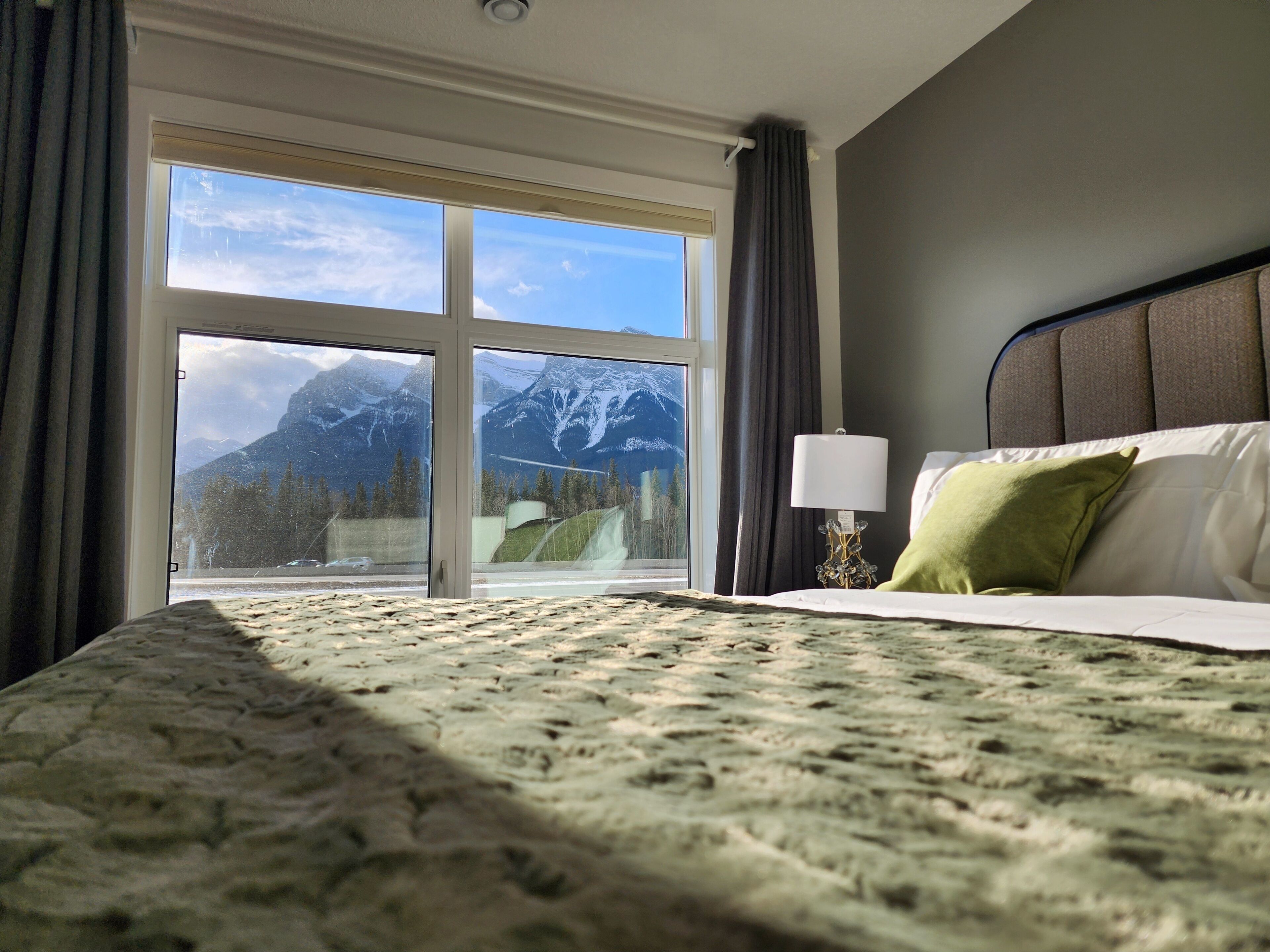 A hotel room with a bed, a nightstand, a lamp, and a view of mountains through a large window.