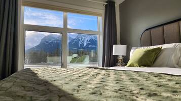 A hotel room with a bed, a nightstand, a lamp, and a view of mountains through a large window.