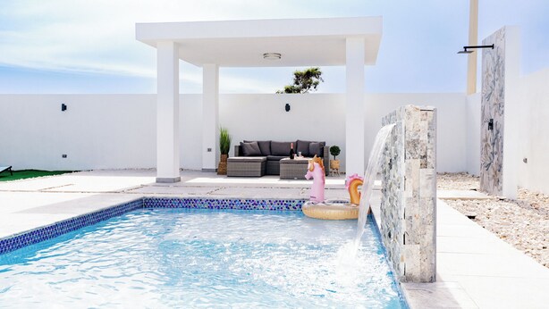 A swimming pool with a stone wall, a covered seating area, and a small waterfall feature. - Boho Style with Private Pool, Cozy 3-bedroom house in beautiful Guayama WiFi, AC (Guayama)