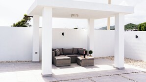 A modern outdoor seating area with a white pergola, cushioned seating, and a small table with a bottle and fruit. - Boho Style with Private Pool, Cozy 3-bedroom house in beautiful Guayama WiFi, AC (Guayama)