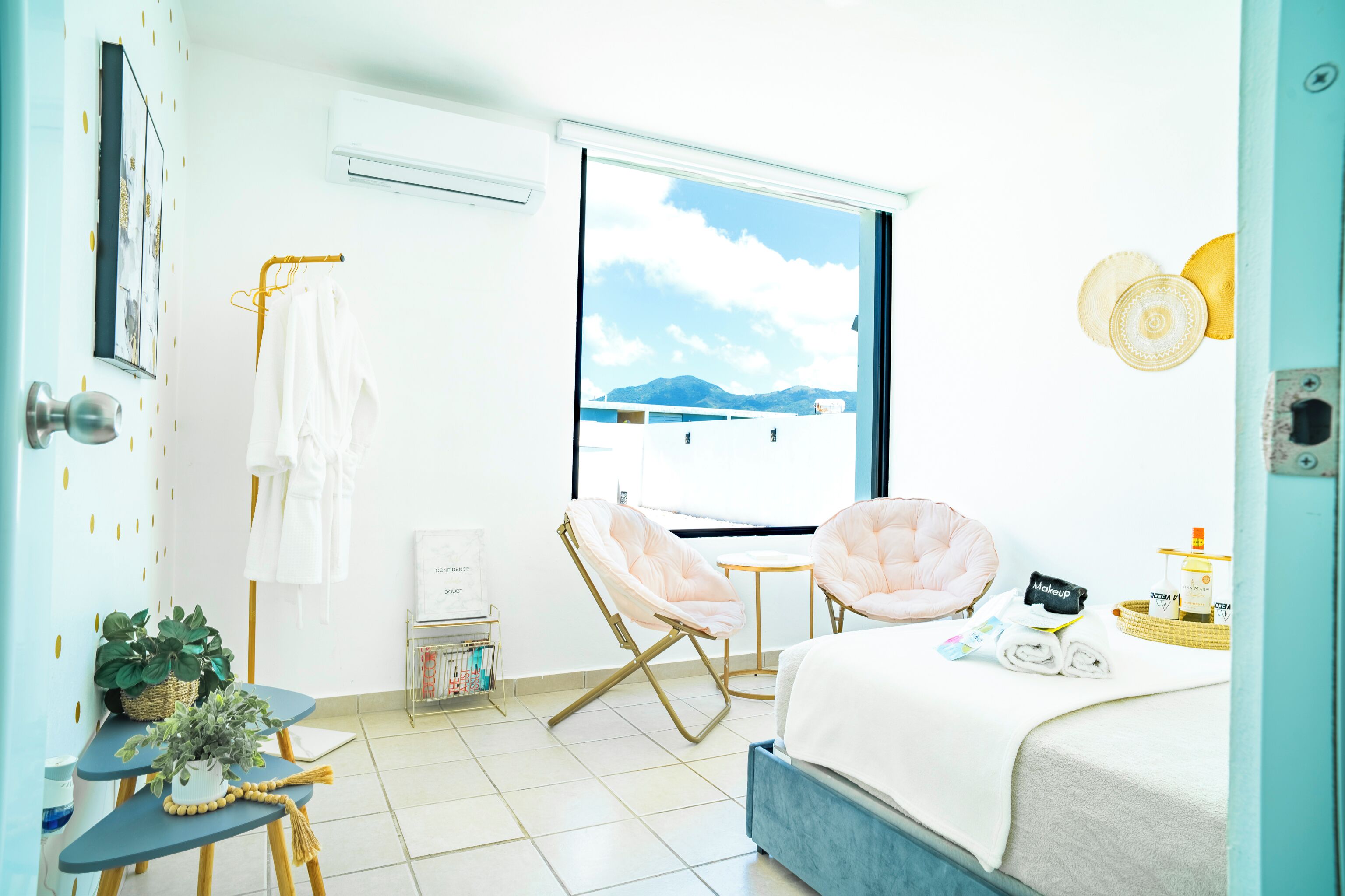 A hotel room with a bed, two chairs, a small table, a window with a view of mountains, and a wall-mounted air conditioner.