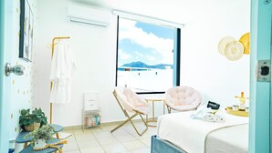 A hotel room with a bed, two chairs, a small table, a window with a view of mountains, and a wall-mounted air conditioner. - Boho Style with Private Pool, Cozy 3-bedroom house in beautiful Guayama WiFi, AC (Guayama)