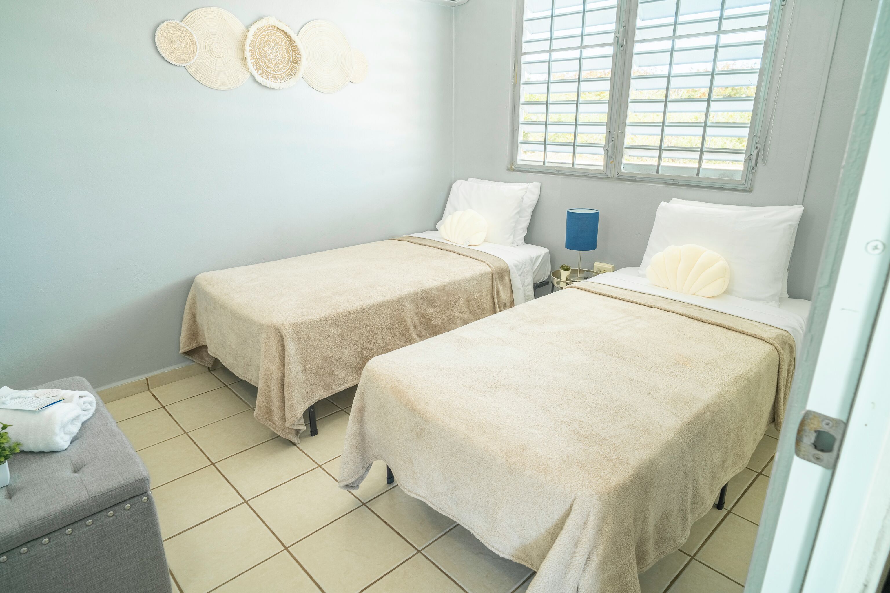 Une chambre avec deux lits, une petite table sur laquelle se trouvent une serviette et une plante, une fenêtre avec des stores et un mur décoré d’assiettes.