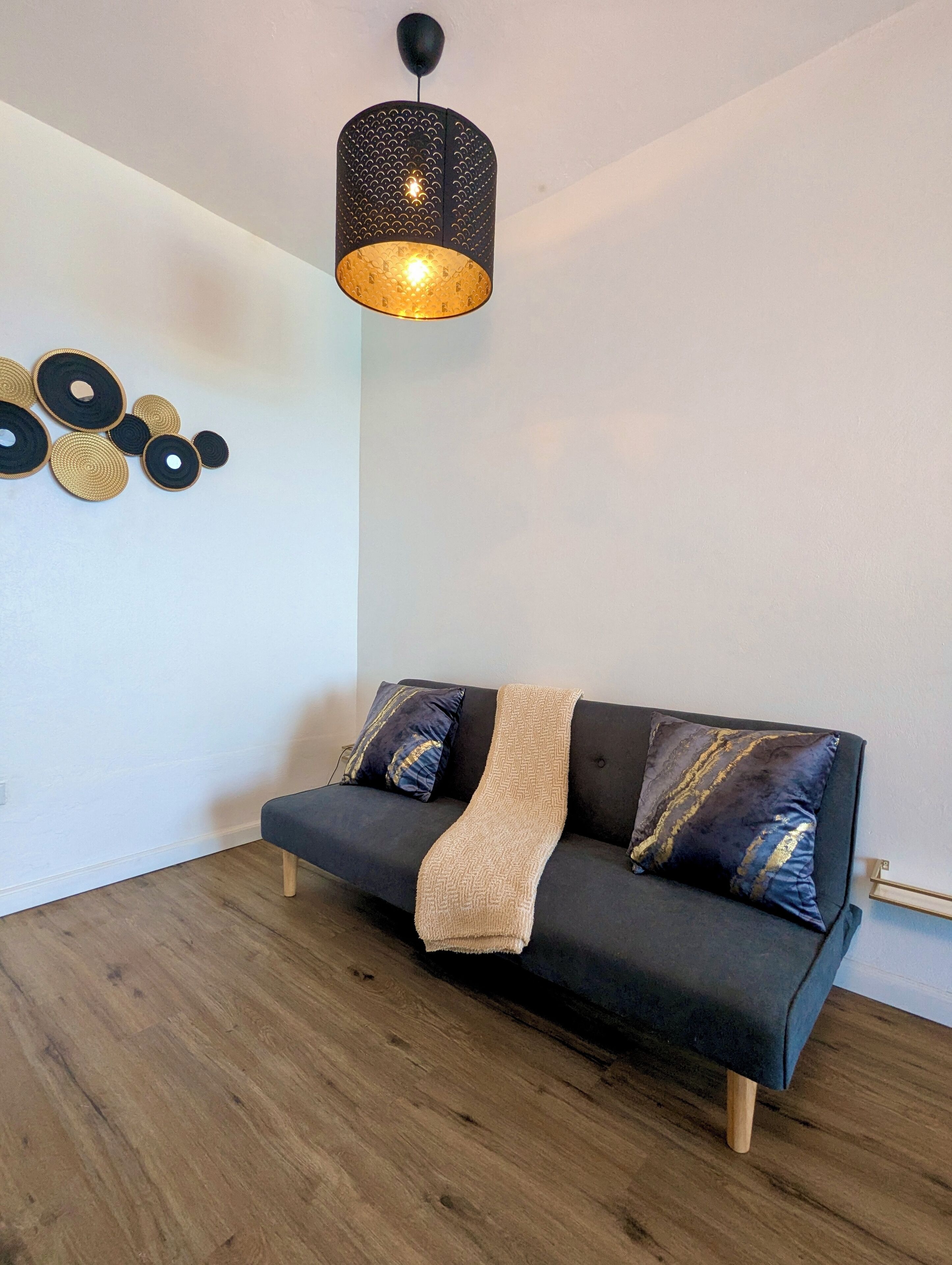 A modern living room with a dark grey sofa, two patterned throw pillows, and a beige throw blanket. A unique black and gold pendant light hangs from the ceiling.