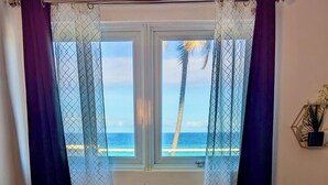 A room with a bed, a window with curtains, a wooden floor, and a view of the beach. - Modern Oceanfront Apartment  (Arecibo)