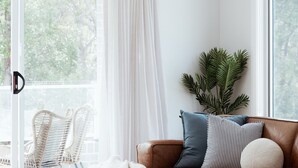 Una habitación con un ventanal grande, una silla de mimbre, un sofá de cuero marrón con almohadones a rayas y una planta verde.