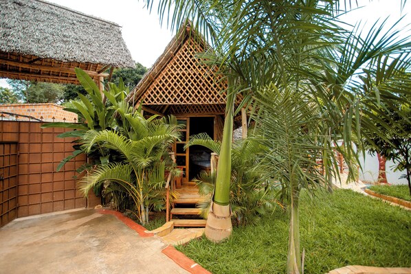 Una estructura con techo de paja y entrada de celosía de madera, rodeada de abundante vegetación y un sendero de concreto que conduce a ella.