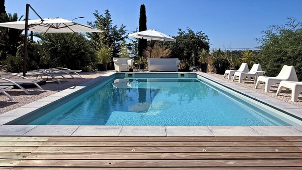 Une piscine rectangulaire à l’eau d’un bleu cristallin, entourée de chaises longues blanches et de parasols, installée sur une terrasse en bois.