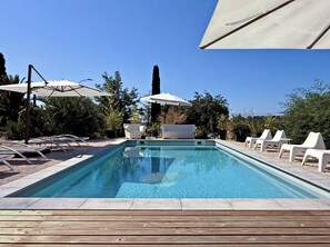 Una piscina rectangular con agua azul cristalina, rodeada de sillas de descanso y sombrillas, ubicada sobre una terraza de madera.