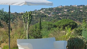 Un espace extérieur aménagé avec des sièges blancs et un foyer, entouré de gravier et de plantes, offrant une vue sur une colline parsemée de maisons.