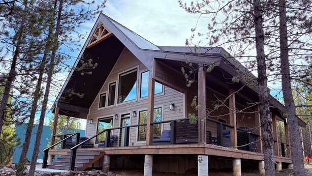 A modern cabin with a large deck, multiple windows, and a sloped roof, surrounded by trees.