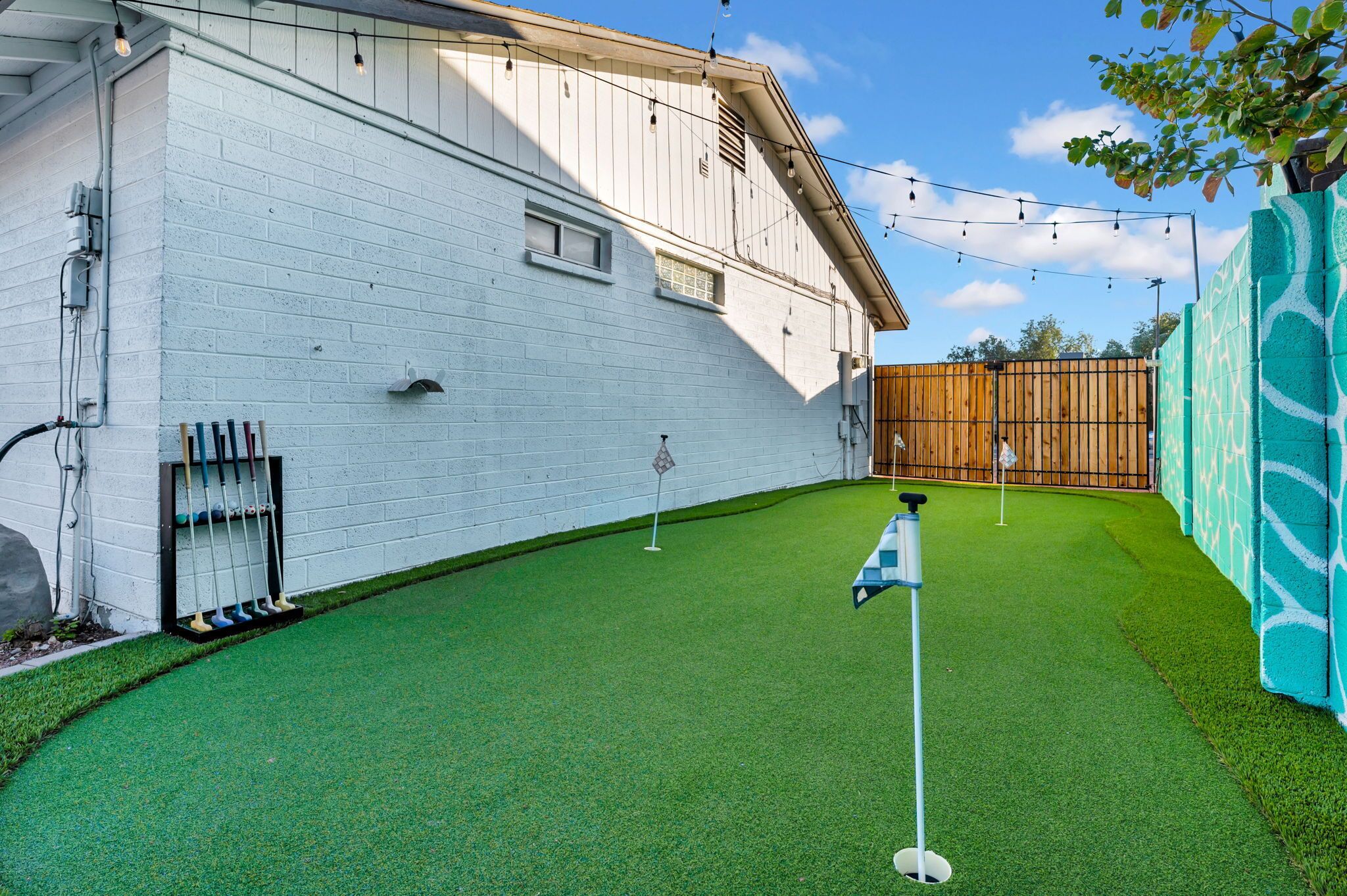 A white building with a brick wall, a wooden fence, and a green artificial turf area with golf flags.