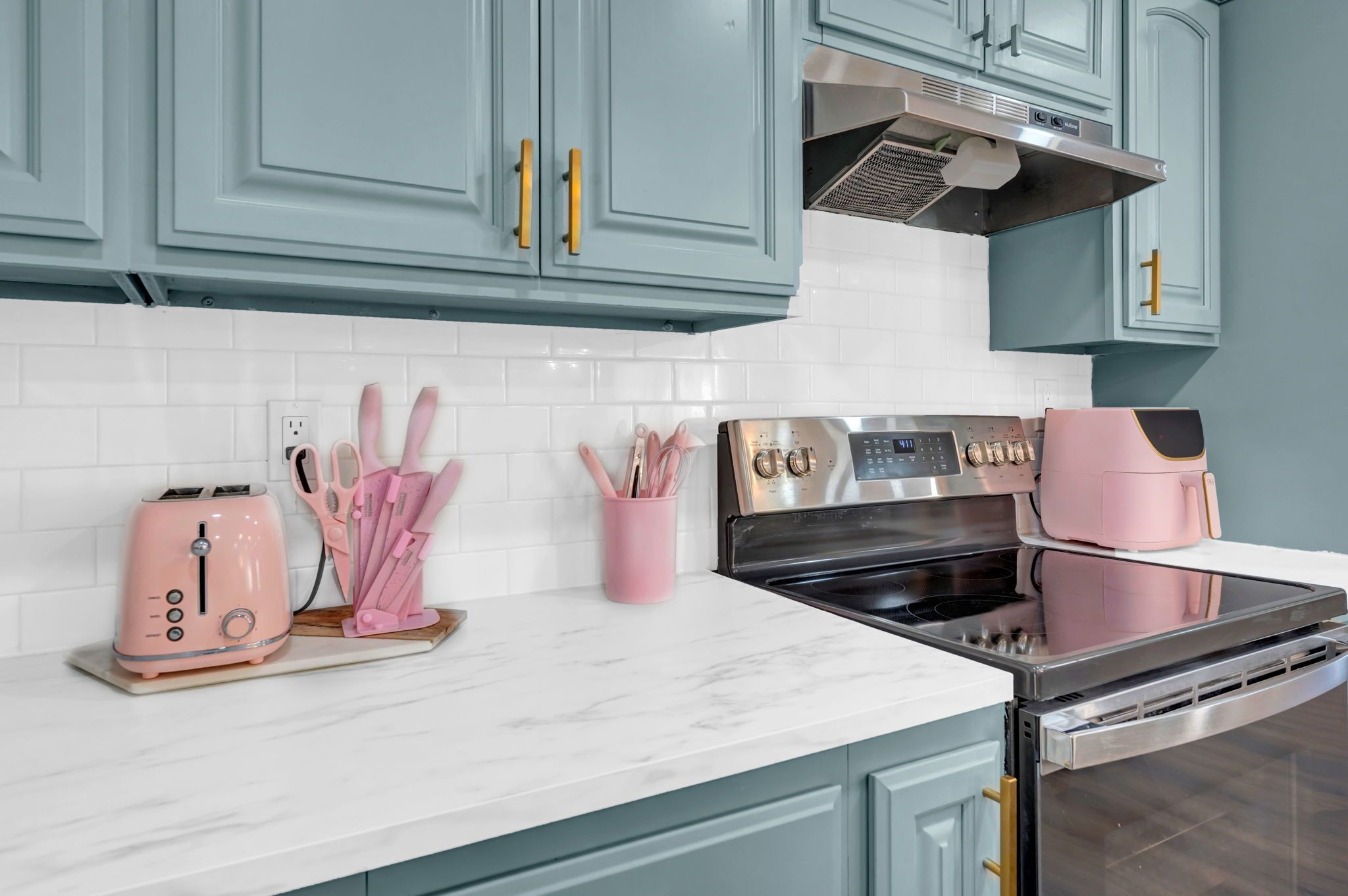 A kitchen with teal cabinets, a white tiled backsplash, a pink toaster, a pink coffee maker, and a stainless steel oven.