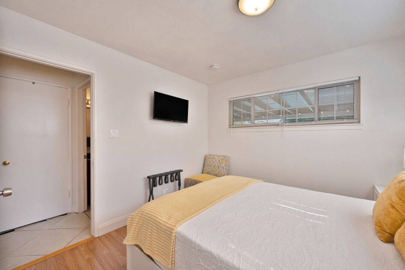 A bedroom with a bed, a TV mounted on the wall, a window with blinds, and a door.