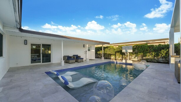 Zona de piscina exterior moderna con cielo azul despejado, espacio de descanso techado con cojines y una canasta de baloncesto.