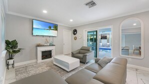 Una sala de estar moderna con una televisión de pantalla plana, una chimenea, un sofá, un sillón y vistas a una piscina a través de puertas de vidrio.