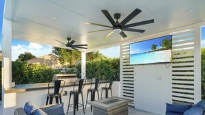 Un área de estar al aire libre techada con mobiliario de mimbre, ventiladores de techo y una televisión que muestra una escena de playa.