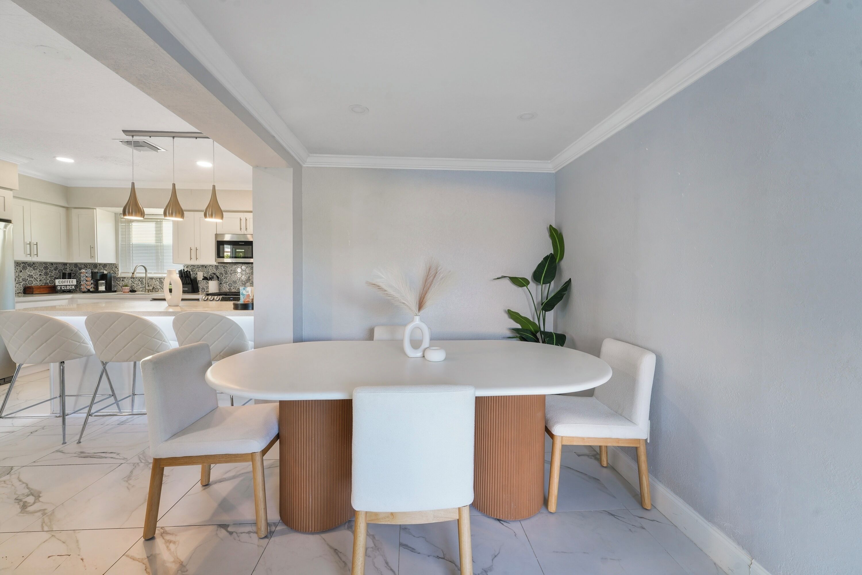Un coin repas moderne avec une table ovale blanche, quatre chaises blanches et un vase contenant des plumes d’herbe de la pampa.