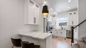 A modern kitchen with white cabinetry, a marble countertop, a stainless steel refrigerator, and a black pendant light. - Powder Mountain Ski Retreat, an art-inspired townhome in the heart of Eden (Eden)