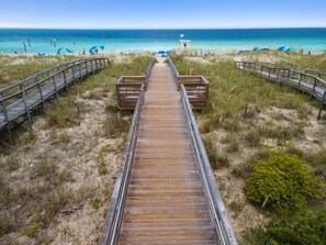 A wooden boardwalk leading to a beach with clear blue water and a lifeguard tower in the distance.