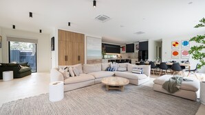En moderne stue med en hjørnesofa, et sofabord og et spiseområde med bord og stole. Der er et køkken i baggrunden med udsigt til det fri.