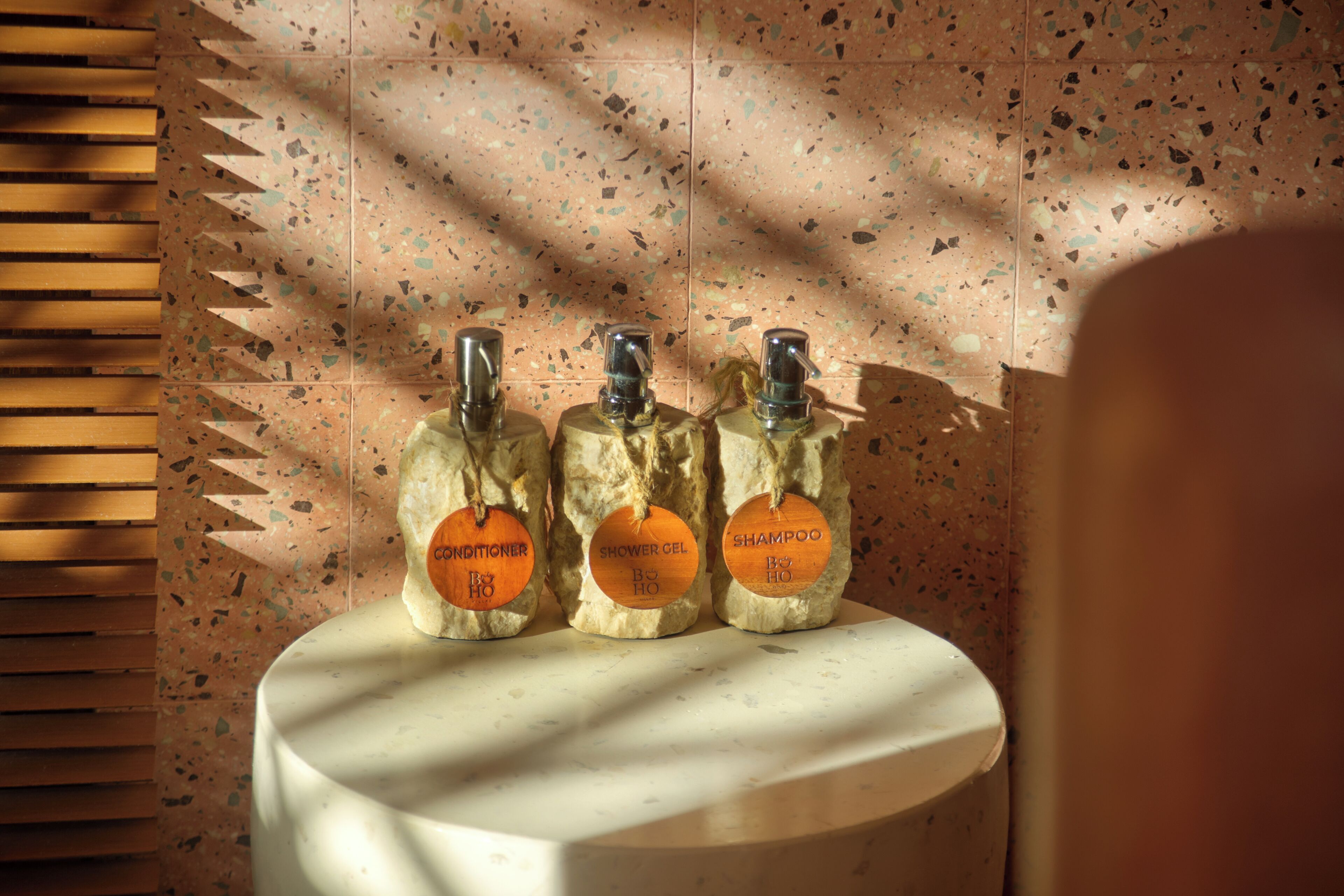 Three toiletry bottles with wooden labels on a white pedestal against a speckled wall.