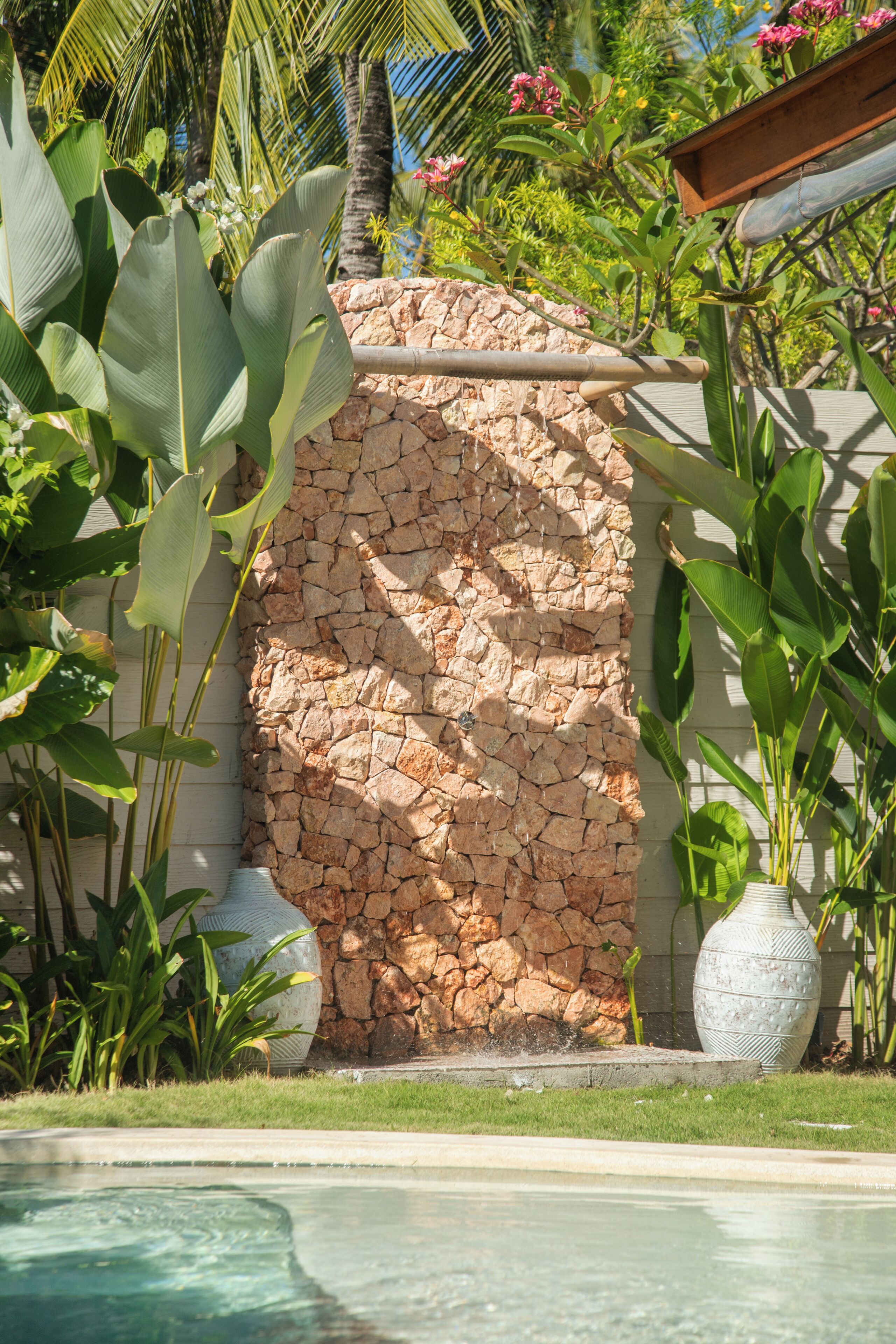 A stone wall with a water feature in front, surrounded by tropical plants and a clear pool.