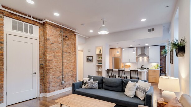 A modern living room with a brick wall, a sofa, a coffee table, and a dining area in the background. - Modern Condo in the heart of Over-The-Rhine (Cincinnati)