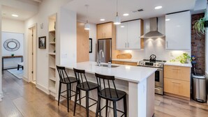 A modern kitchen with a central island, bar stools, stainless steel appliances, and a hanging plant. - Modern Condo in the heart of Over-The-Rhine (Cincinnati)